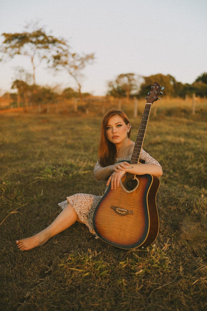 menu-02 A woman sits on a grassy field, embracing an acoustic guitar at sunset, exuding calmness and musical passion.