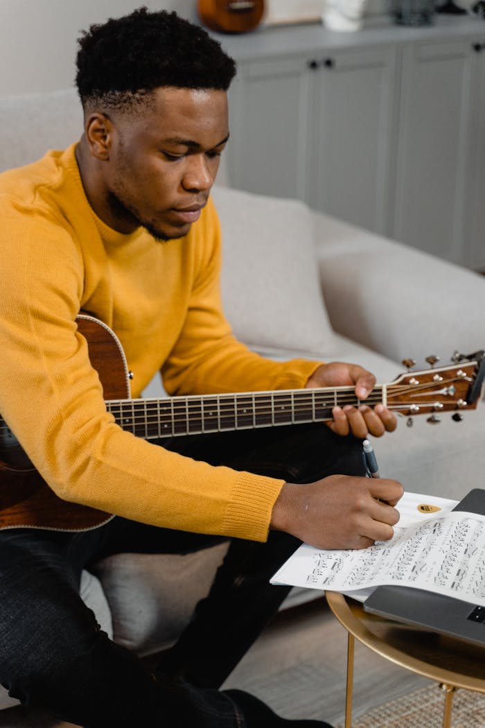 menu-01 African American man writing sheet music while holding a guitar indoors.