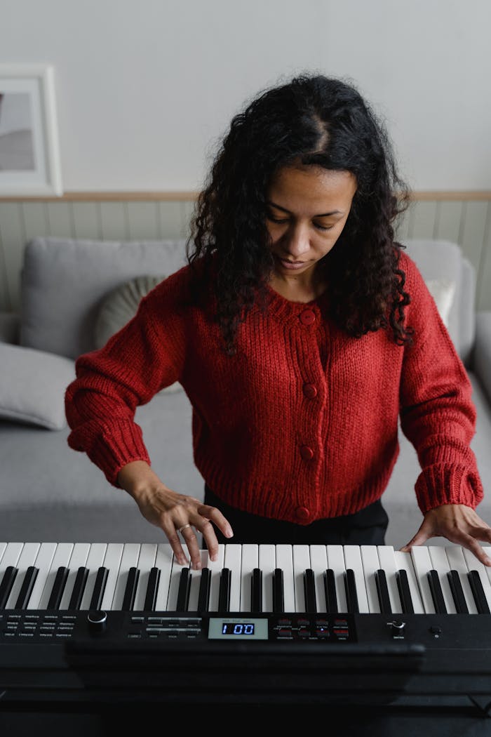 menu-08 Young woman in a red sweater plays a keyboard indoors, focusing on music.