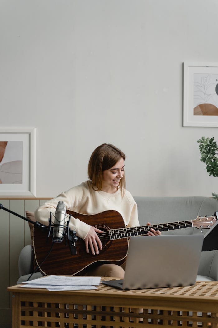 menu-09 Caucasian woman playing guitar while smiling and sitting on a couch with a laptop nearby.
