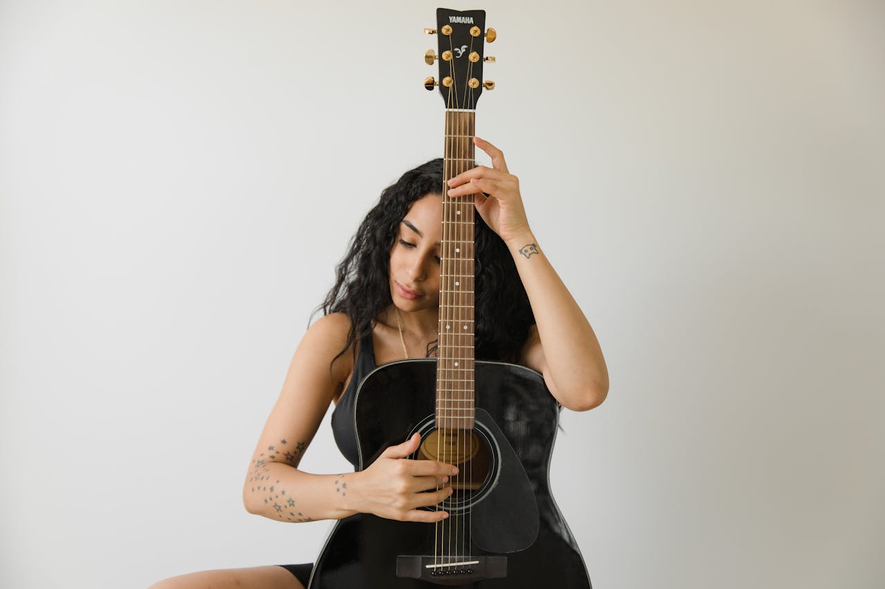 menu-14 Woman with curly hair plays a black acoustic guitar indoors, focusing on her music.